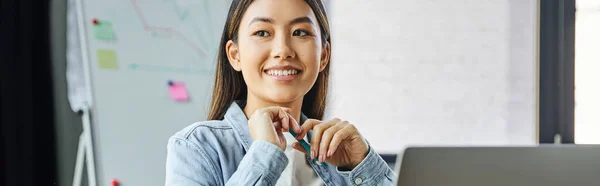 Задоволена азіатська жінка з брюнеткою волоссям і щасливою посмішкою тримає ручку і дивиться геть, сидячи в сучасному офісі, молода успішна бізнес-леді, банер — стокове фото