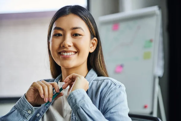 Portrait of young and charming asian businesswoman with brunette hair and cheerful smile holding pen and looking at camera in modern office, professional headshot — Stock Photo