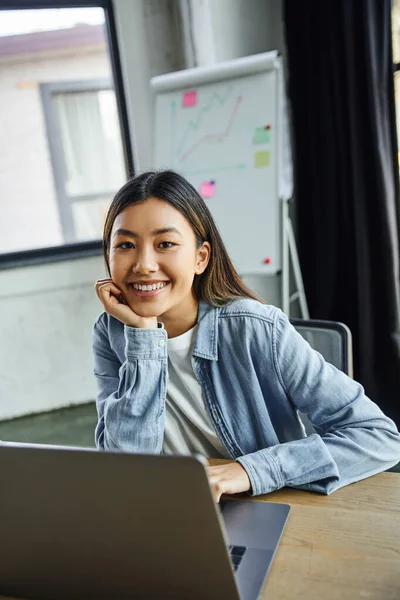 Молодая и довольная азиатская бизнесвумен с брюнетками, в джинсовой рубашке, смотрящая в камеру возле ноутбука и флип-чарт с графиками на размытом фоне в современном офисе — стоковое фото