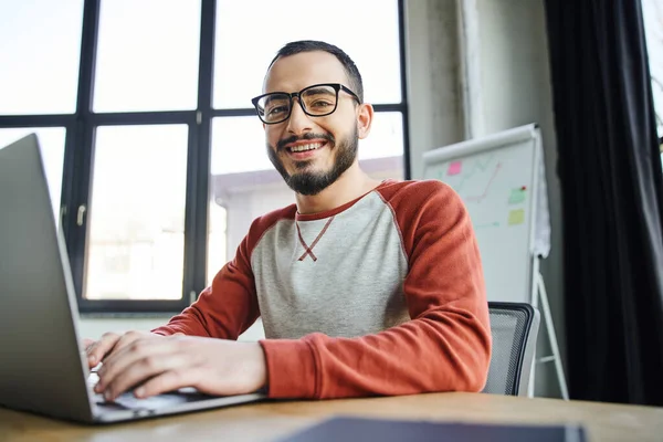 Jovem e sorridente barbudo empresário em óculos digitando no laptop e olhando para a câmera na frente de flip chart com gráficos sobre fundo borrado no escritório contemporâneo — Fotografia de Stock