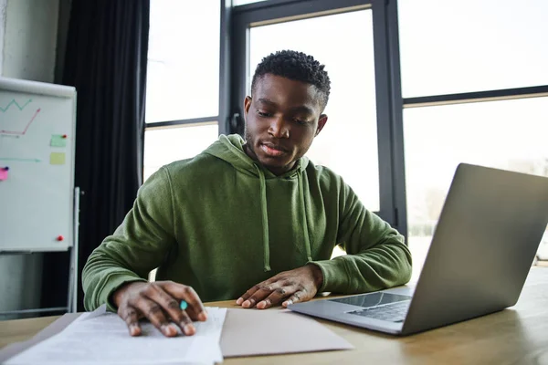 Jovem e elegante empresário afro-americano sentado no local de trabalho e trabalhando com documentos perto de laptop e flip chart em fundo turvo no escritório, jovens ambiciosos e orientados para a carreira — Fotografia de Stock