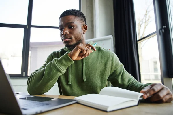 Молодий і красивий афроамериканський бізнесмен в зеленому стильному светрі дивиться на ноутбук біля ноутбука, сидячи на робочому місці і працюючи на розмитому передньому плані в сучасному офісі — стокове фото