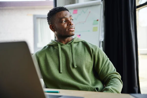 Молодий афроамериканський підприємець у зеленій шубці сидить і дивиться далеко, плануючи стартап біля фліп-чарту з бізнес-аналітикою в сучасному офісі, професійний розвиток — стокове фото