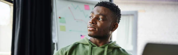 Portrait of youthful and thoughtful african american entrepreneur in green hoodie looking away near flip chart with business analytics in modern office, professional development, banner — Stock Photo