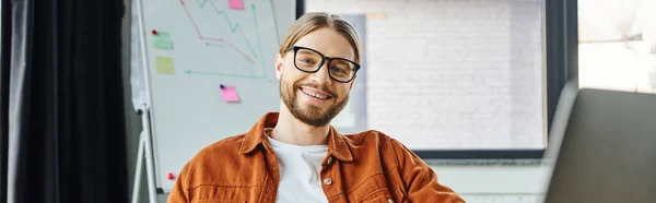 Hombre de negocios barbudo lleno de alegría en gafas y camisa de moda, con sonrisa radiante, mirando a la cámara mientras está sentado frente al rotafolio sobre fondo borroso en la oficina moderna, pancarta - foto de stock