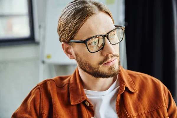 Portrait of thoughtful and bearded businessman in eyeglasses and trendy shirt looking away in contemporary office, thinking and planning startup project, blurred background — Stock Photo