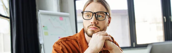 Portrait of serious and thoughtful bearded businessman in eyeglasses and trendy shirt holding hands under chin and looking away near flip chart on blurred background in office, banner — Stock Photo