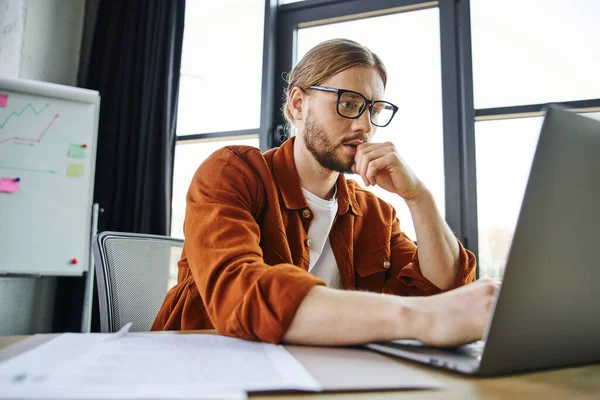Concentrated bearded businessman in eyeglasses and trendy shirt working in office, thinking near laptop, documents and flip chart with analytics on blurred background — Stock Photo