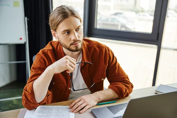 Вдумчивый бородатый бизнесмен в модной рубашке сидит рядом с ноутбуком и документами на рабочем столе, держит очки и смотрит в сторону, серьезный и стильный предприниматель работает в современном офисе — стоковое фото