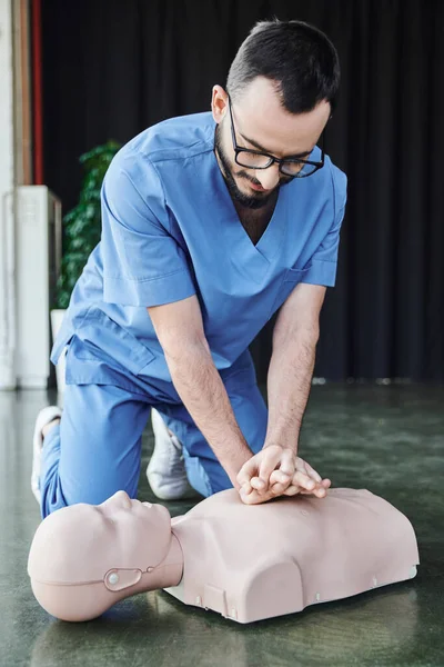 Young medical instructor in eyeglasses and uniform doing chest compressions on CPR manikin on floor in training room, effective life-saving skills and emergency preparedness concept — Stock Photo