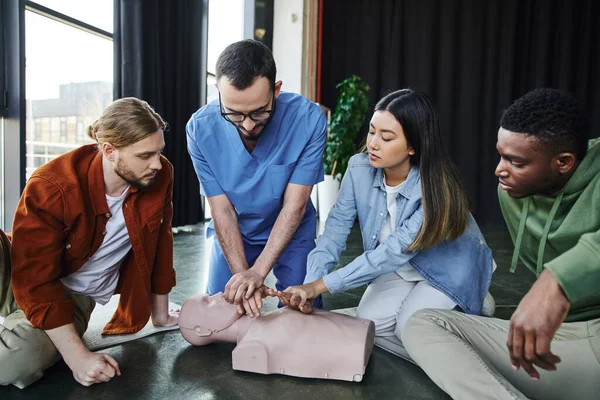 First aid training, medical instructor showing cardiopulmonary resuscitation on CPR manikin near multiethnic participants in training room, effective life-saving skills and techniques concept — Stock Photo
