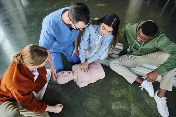 First aid seminar, cardiopulmonary resuscitation training, high angle view of young asian woman doing chest compressions on CPR manikin near medical instructor and multiethnic team — Stock Photo