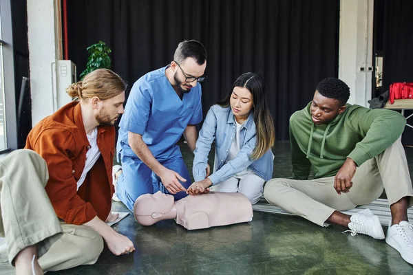 Young asian woman practicing life-saving skills by doing chest compressions on CPR manikin near multiethnic team and medical instructor, cardiopulmonary resuscitation, first aid training seminar — Stock Photo