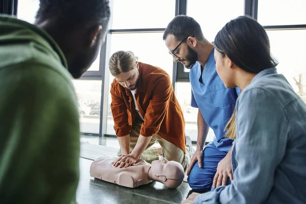 Cardiopulmonary resuscitation, young man doing chest compressions on CPR manikin during hands-on learning on first aid training seminar near medical instructor and multiethnic participants — Stock Photo