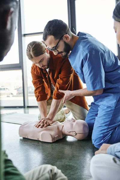 First aid training seminar, professional paramedic helping young man doing chest compressions on CPR manikin near multiethnic participants, effective life-saving skills and techniques concept — Stock Photo