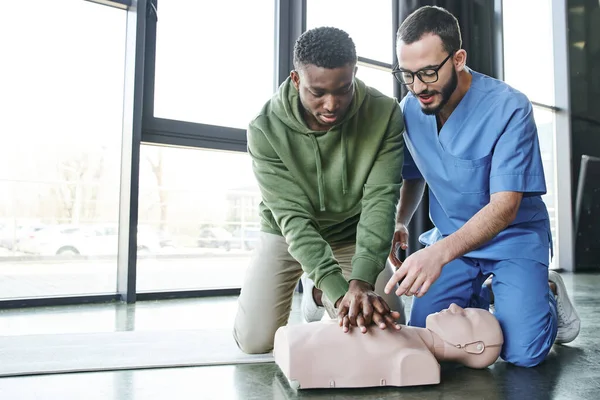 Medizinischer Instruktor in Uniform und Brille, der auf eine CPR-Schaufensterpuppe zeigt, während ein afrikanisch-amerikanischer Mann Brustkompressionen macht, effektive lebensrettende Fähigkeiten und Notfallvorsorgekonzept — Stockfoto