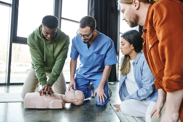 Junge multiethnische Menschen und medizinischer Ausbilder, die einen afrikanisch-amerikanischen Mann bei Brustkompressionen an CPR-Schaufensterpuppen, Herz-Lungen-Wiederbelebung, Erste-Hilfe-Schulung beobachten — Stockfoto