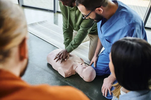 African american man practicing chest compressions and cardiopulmonary resuscitation on CPR manikin near medical instructor and young participants of first aid seminar in training room — Stock Photo