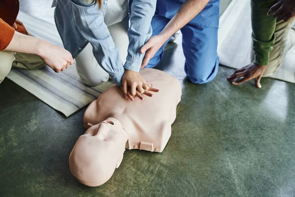 Cropped view of paramedic assisting young woman practicing chest compressions on CPR manikin near multiethnic participants of first aid training seminar, life-saving skills and techniques concept — Stock Photo
