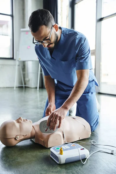 First aid training seminar, young medical instructor in uniform and eyeglasses applying defibrillator pads on CPR manikin, cardiac resuscitation, health care and life-saving techniques concept — Stock Photo
