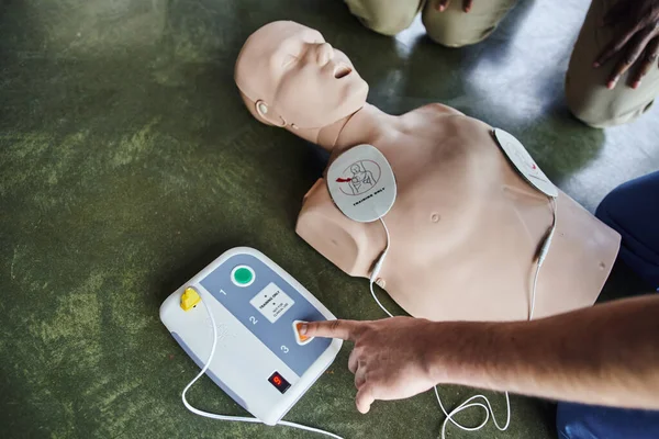 Partial view of professional medical instructor operating defibrillator on CPR manikin near young participants of first aid seminar, high angle view, health care and life-saving techniques concept — Stock Photo