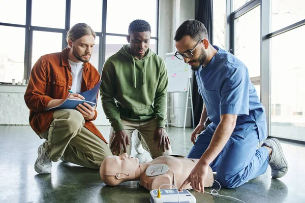 Herz-Wiederbelebung, junger Mann schreibt in Notizbuch in der Nähe eines afrikanisch-amerikanischen Teilnehmers und medizinischer Instruktor bedient Defibrillator an CPR-Schaufensterpuppe während eines Erste-Hilfe-Trainings — Stockfoto