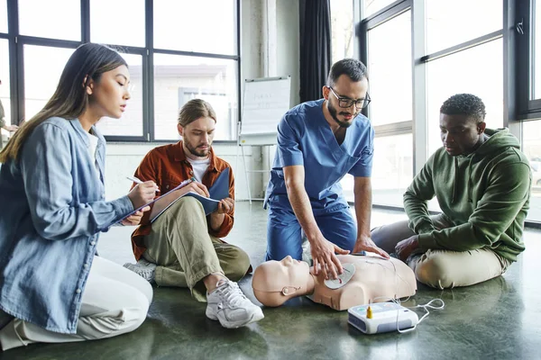 Cardiac resuscitation skills, healthcare worker applying defibrillator pads on CPR manikin near young diverse and multiethnic group, health care and life-saving techniques concept — Stock Photo