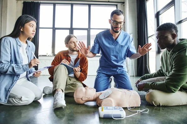 Geste paramédical professionnel et parler aux participants multiethniques près du mannequin et du défibrillateur de la RCR pendant le séminaire de premiers soins dans la salle de formation, le concept des soins de santé et des techniques de sauvetage — Photo de stock