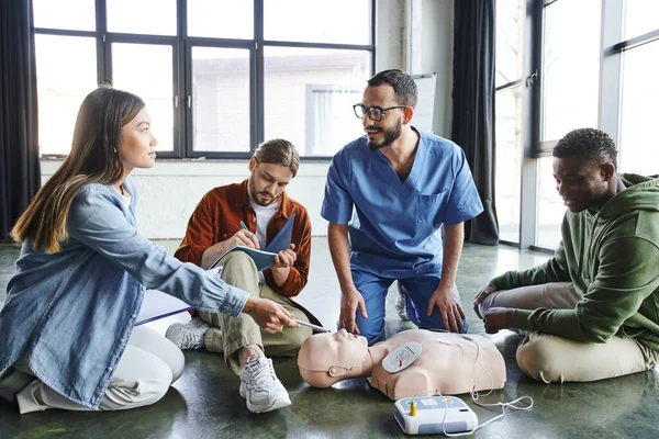 First aid hands-on learning, asian woman with clipboard pointing with pen at CPR manikin with defibrillator near paramedic and multicultural men, health care and life-saving techniques concept — Stock Photo