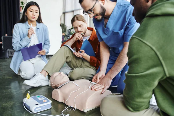 First aid seminar, medical instructor applying defibrillator pads on CPR manikin near multiethnic team with clipboard and notebook in training room, health care and life-saving techniques concept — Stock Photo