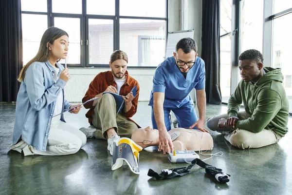 Professional paramedic showing cardiac resuscitation techniques with CPR manikin and defibrillator to multicultural participants of first aid seminar, life-saving skills and emergency care concept — Stock Photo
