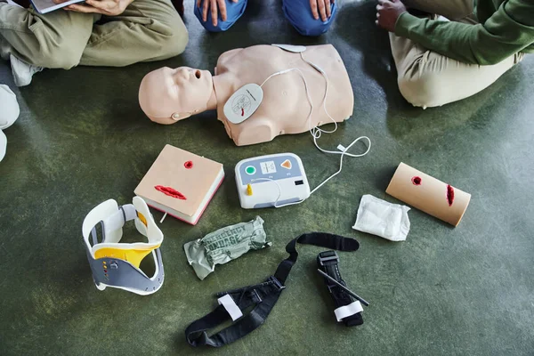 Cropped view of multiethnic participants of first aid seminar near CPR manikin, defibrillator, wound care simulators, bandages, compression tourniquets and neck brace in training room, top view — Stock Photo