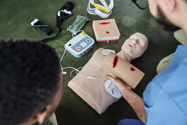 Vue partielle du professionnel paramédical tenant simulateur de soins des plaies près de l'homme afro-américain, mannequin CPR avec défibrillateur et équipement médical, concept de préparation aux situations d'urgence, vue de dessus — Photo de stock