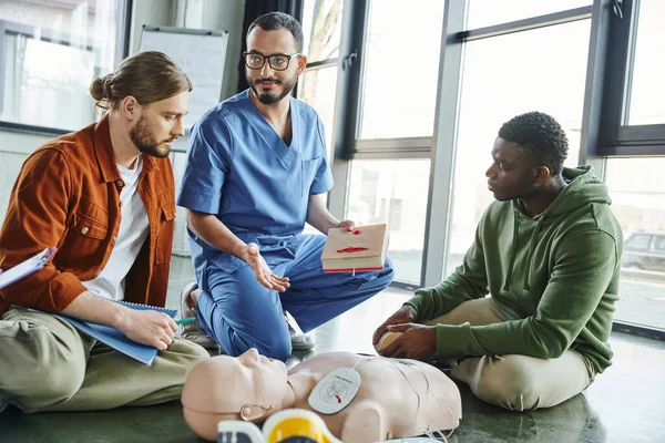 Smiling healthcare worker in eyeglasses and uniform holding wound care simulator and pointing at CPR manikin near multicultural participants of medical seminar, effective life-saving skills concept — Stock Photo