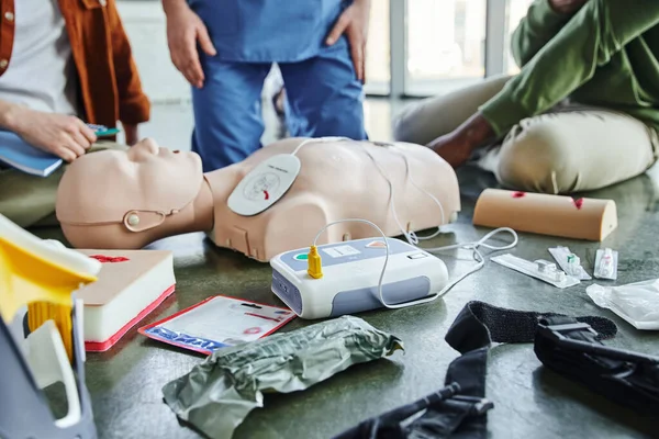 CPR manikin, defibrillator, wound care simulators, compression tourniquet, bandages and syringes near instructor and multiethnic participants of first aid seminar, emergency preparedness concept — Stock Photo