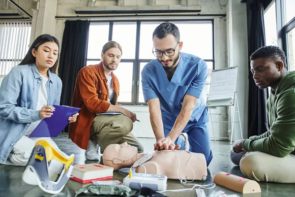 Young asian woman with clipboard looking at paramedic doing chest compressions on CPR manikin near medical equipment and multiethnic participants of first aid seminar in training room — Stock Photo
