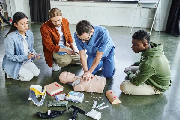 First aid seminar, multiethnic team looking at medical instructor doing chest compressions on CPR manikin near defibrillator, wound care simulators, neck brace, bandages and compression tourniquets — Stock Photo