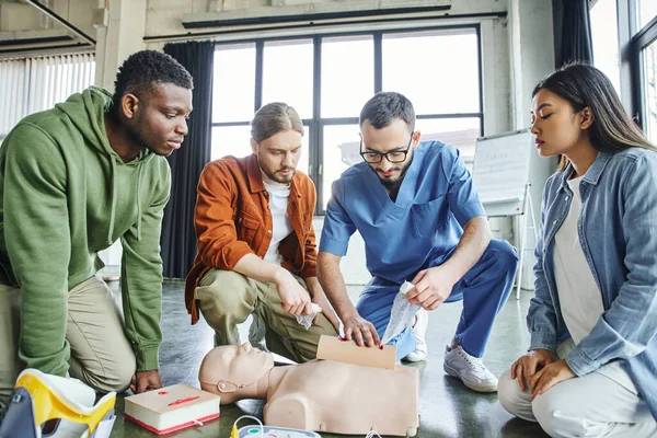 First aid seminar, hands-on learning, multicultural team looking at medical instructor tamponing wound on simulator with bandage near medical equipment, life-saving skills concept — Stock Photo