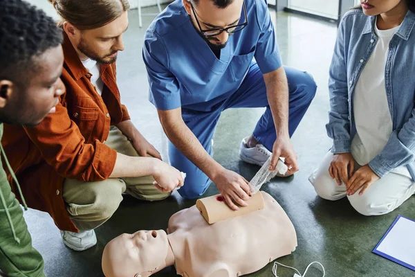 High angle view of professional paramedic tamponing wound on simulator with bandage during first aid seminar with diverse multiethnic group, life-saving skills and emergency response concept — Stock Photo