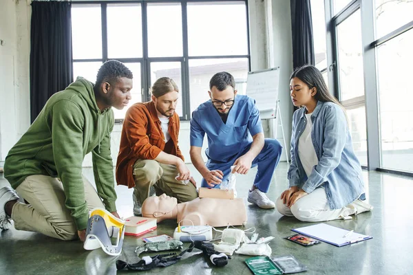 Young and multiethnic participants of medical seminar looking at paramedic tamponing wound on simulator with bandage near medical equipment in training room, life-saving skills concept — Stock Photo