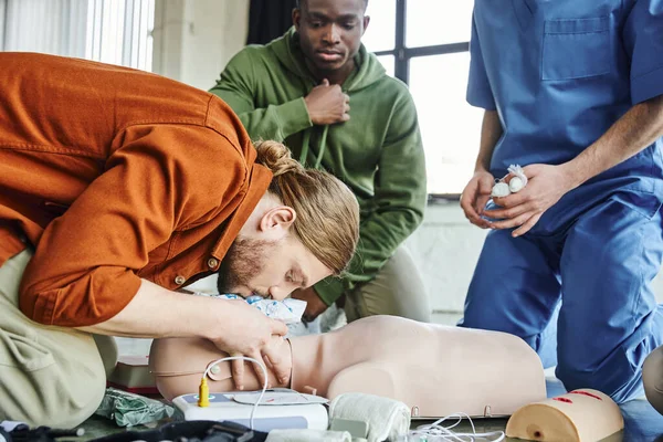 First aid seminar, young man practicing life-saving skills and doing artificial respiration to CPR manikin near defibrillator, medical equipment and african american man with medical instructor — Stock Photo