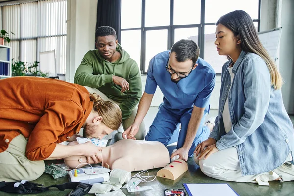 Young man practicing artificial respiration on CPR manikin near defibrillator, medical equipment, multiethnic team and paramedic during hands-on learning on first aid seminar — Stock Photo