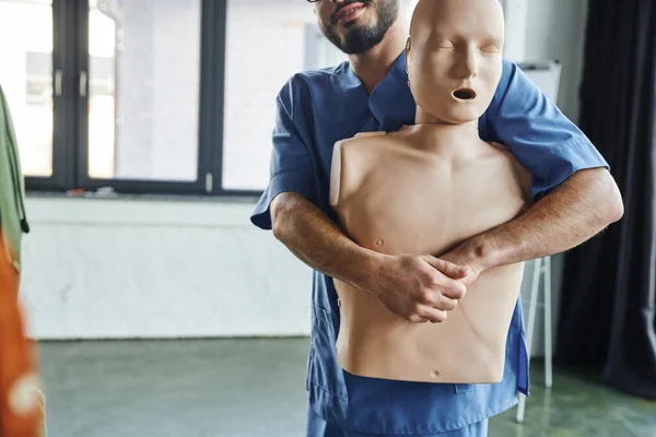 First aid training seminar, partial view of bearded medical instructor in uniform practicing life-saving techniques in case of chocking on CPR manikin, emergency situations preparedness concept — Stock Photo