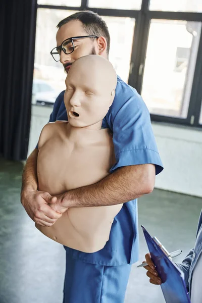 Erste-Hilfe-Seminar, junge professionelle Sanitäter zeigen Rettungstechniken bei Erstickung an CPR-Schaufensterpuppe in der Nähe einer Frau mit Klemmbrett und Stift im Schulungsraum, Vorsorge für Notfälle — Stockfoto