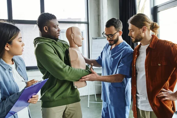 Medical instructor assisting african american man practicing life-saving techniques in case of choking on CPR manikin near multiethnic participants, emergency situations preparedness concept — Stock Photo