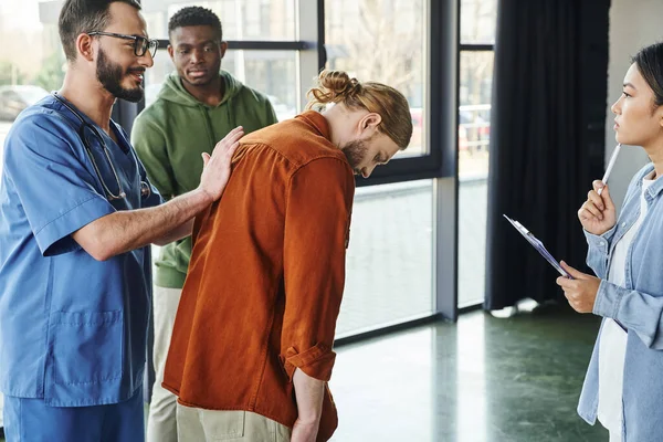 First aid seminar, positive medical instructor in eyeglasses and uniform pushing back of man while showing life-saving techniques in case of choking, emergency situations preparedness concept — Stock Photo