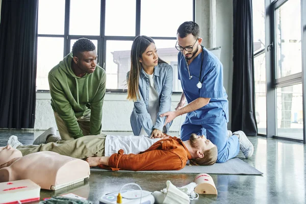 African american man looking at paramedic assisting asian woman doing chest compressions on participant lying near CPR manikin and medical equipment, life-saving skills and techniques concept — Stock Photo
