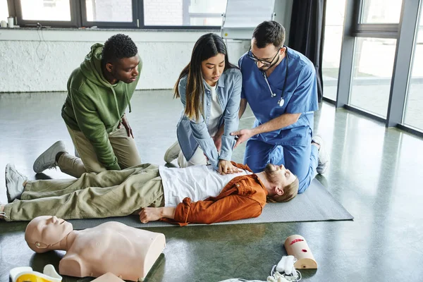 Young asian woman doing chest compressions on man near medical instructor, CPR manikin and african american participant in first aid training seminar, effective life-saving skills concept — Stock Photo