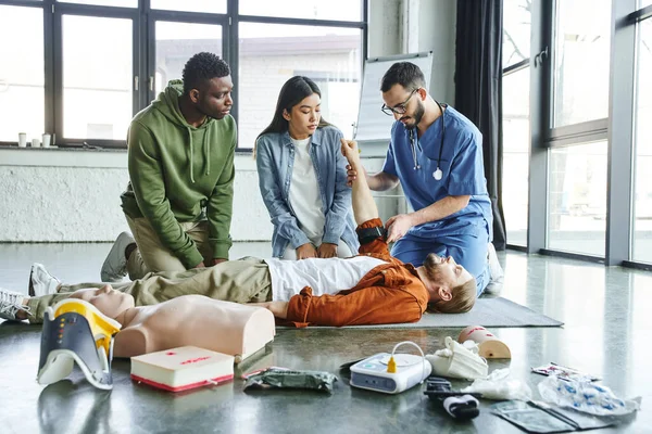 Medical instructor applying compression tourniquet on arm of seminar participant near asian woman, african american man and medical equipment in training room, emergency situations response concept — Stock Photo