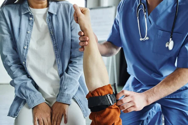 Vue recadrée d'un ambulancier paramédical professionnel appliquant un garrot de compression sur le bras d'un homme près d'une jeune femme lors d'un séminaire de premiers soins en salle d'entraînement, concept de réponse aux situations d'urgence — Photo de stock